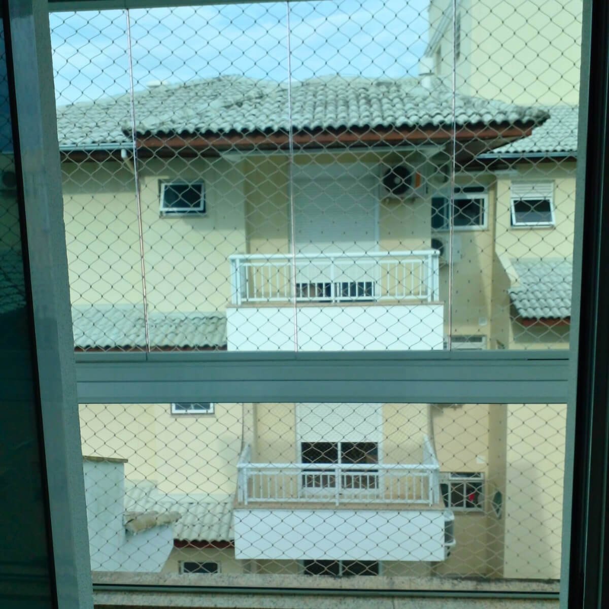 Vista interna de um apartamento através de uma grande janela envidraçada protegida por rede, mostrando o prédio vizinho.