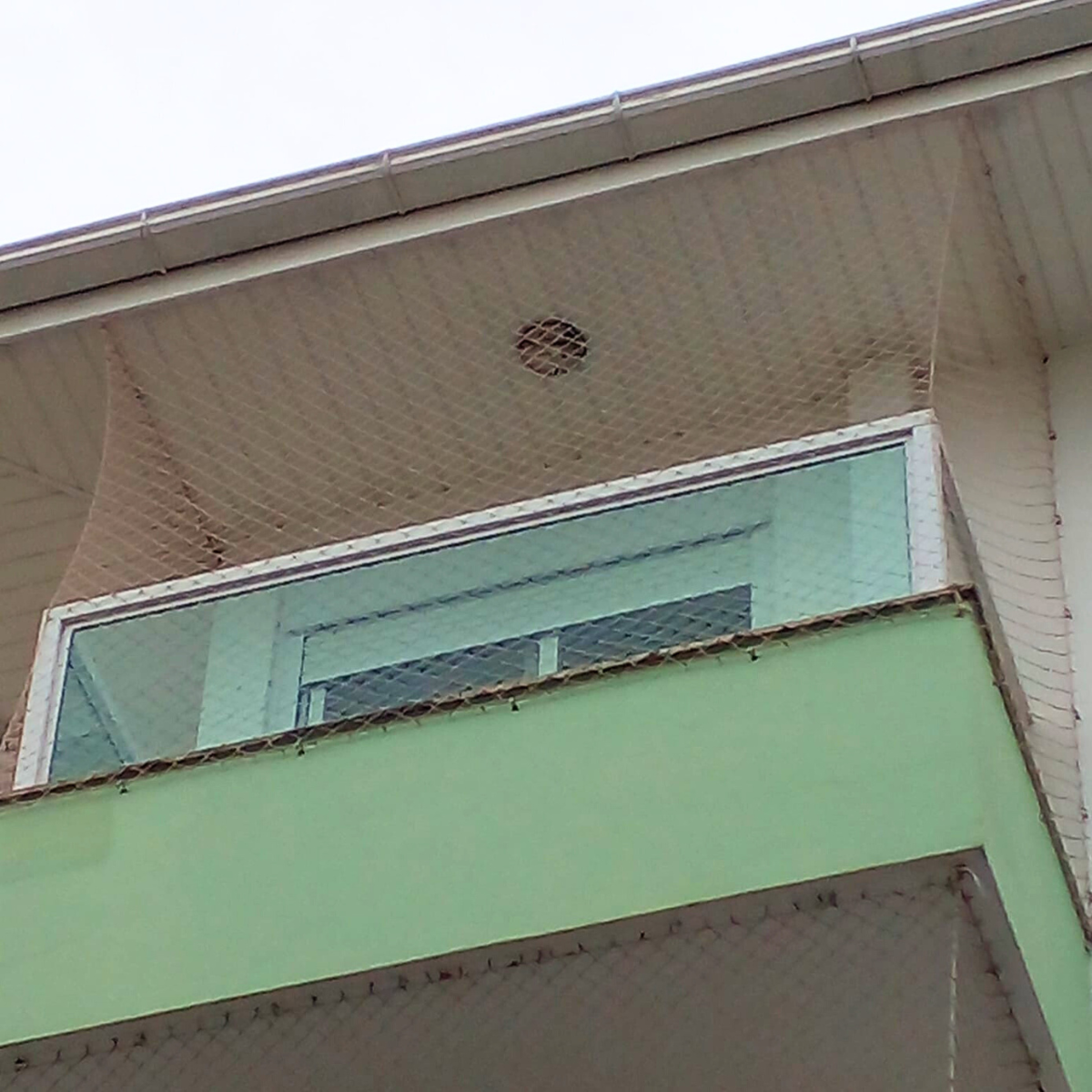 Vista de baixo para cima de uma sacada com frente verde e vidro, mostrando o fechamento total do vão com rede de proteção até o teto (beiral).