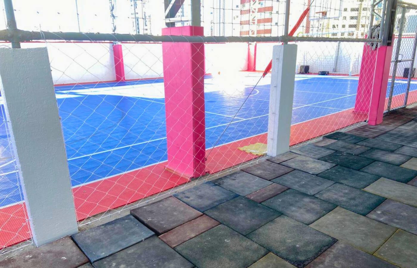 Quadra poliesportiva com piso azul e vermelho e pilares rosa, totalmente cercada por rede de proteção branca (fio grosso).