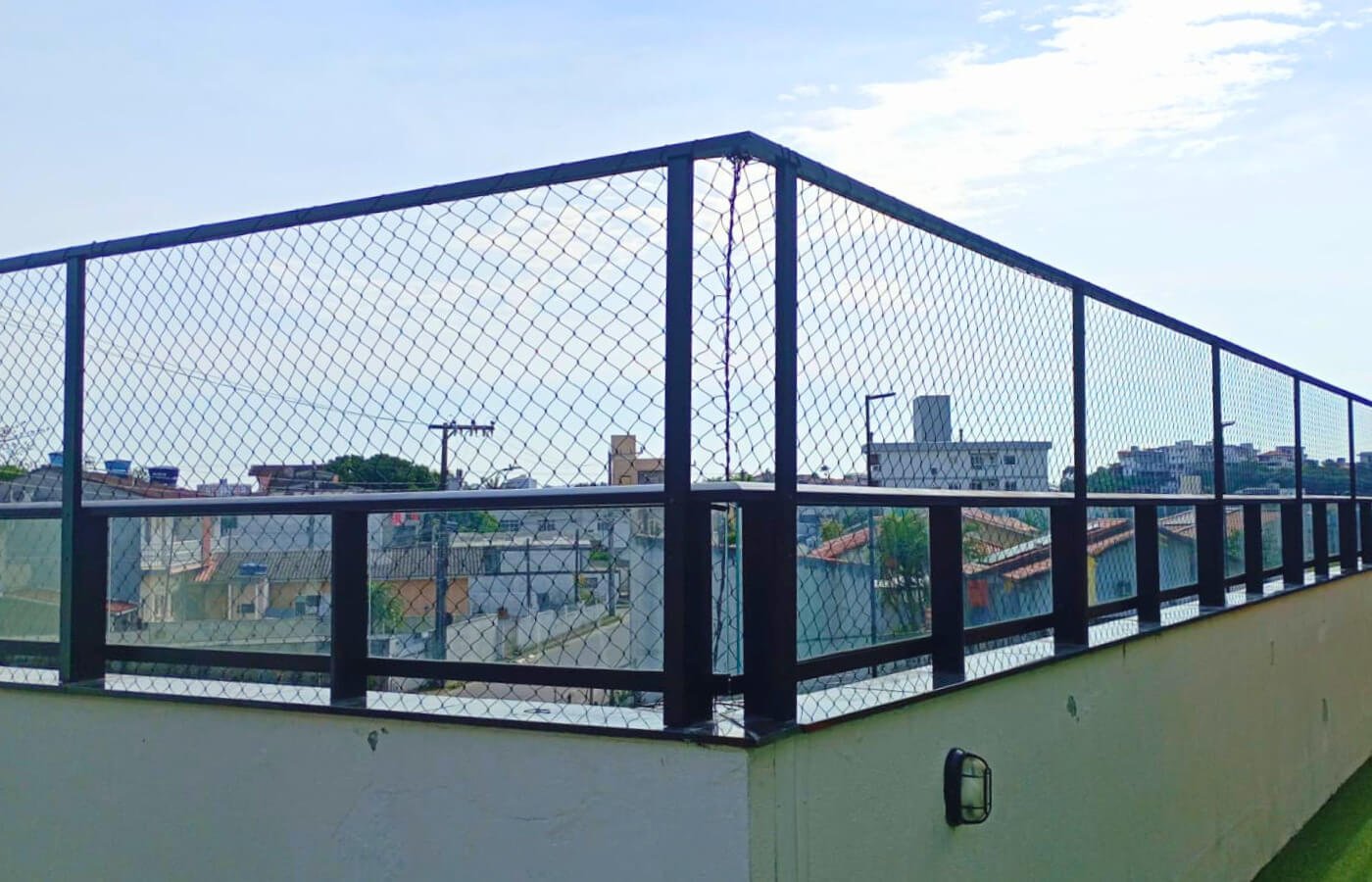 Terraço ou área comum no topo de um prédio, com guarda-corpo preto reforçado por rede de proteção instalada em estrutura metálica.