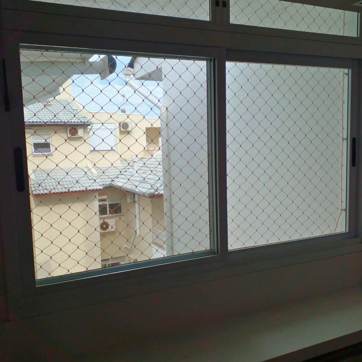 Janela de alumínio branco protegida por rede com vista para fachada interna do prédio vizinho.