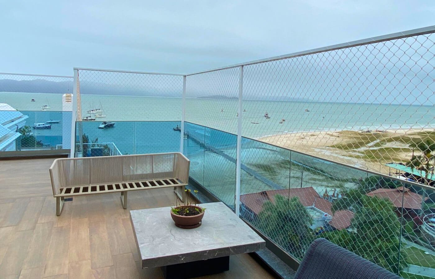 Vista para a praia através de rede de proteção instalada em terraço com mesa de pedra e banco de madeira.