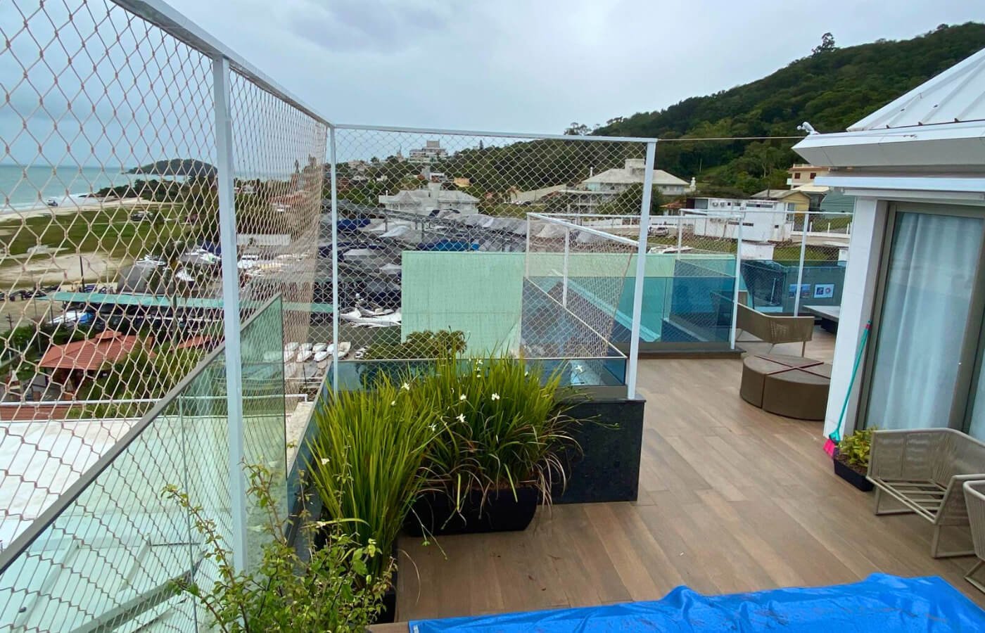 Detalhe lateral de terraço com plantas, mostrando a fixação da rede de proteção sobre o guarda-corpo de vidro.